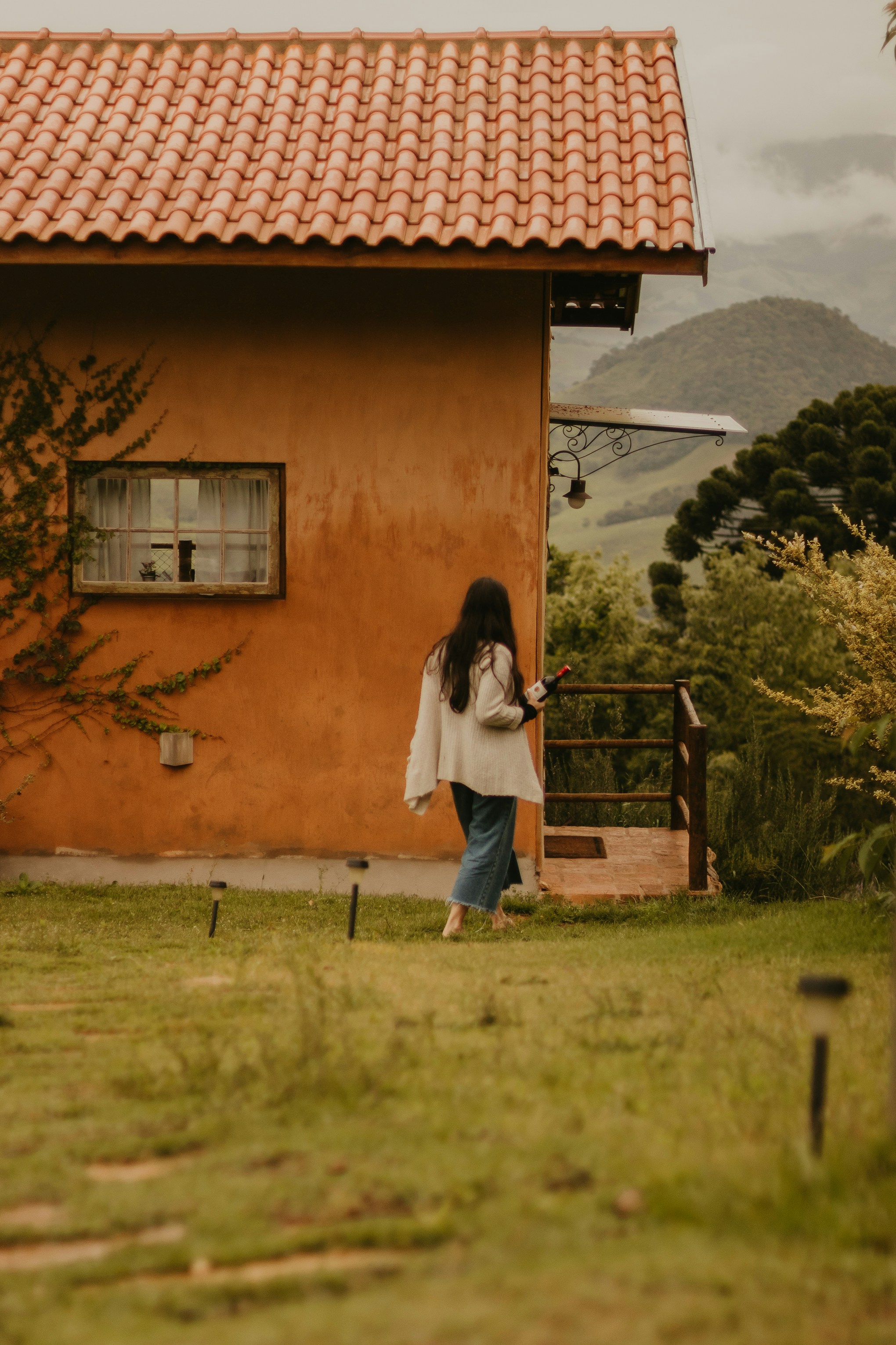 A woman with long dark hair walks barefoot through a grassy yard toward a rustic orange house, holding a bottle of wine, with misty mountains and lush trees in the background